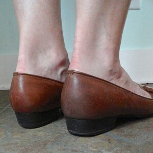 May include: Brown leather loafers with a low heel. The shoes have a classic design with a rounded toe and a small heel. The leather shows signs of wear, with creases and colour variations. The heels are approximately 2.5 cm high.