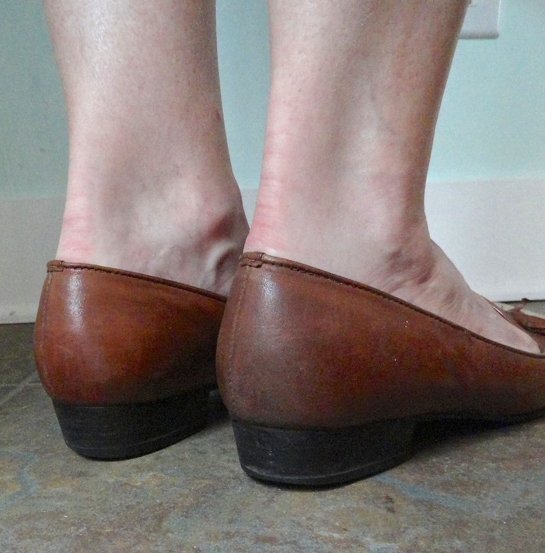 May include: Brown leather loafers with a low heel. The shoes have a classic design with a rounded toe and a small heel. The leather shows signs of wear, with creases and colour variations. The heels are approximately 2.5 cm high.