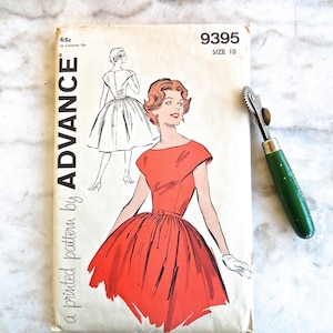 May include: Vintage sewing pattern by Advance, size 10, featuring a red dress illustration. The pattern envelope is cream-colored with text and a drawing of a woman in a dress. A green-handled tracing wheel rests on the side.