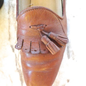 May include: A close-up shot of a brown leather loafer featuring a tassel. The shoe has a rounded toe and a decorative fringe. The leather shows signs of wear, with creases and a slightly glossy finish. A classic shoe style.