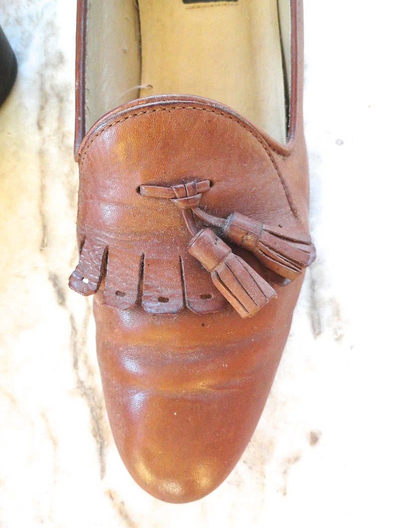 May include: A close-up shot of a brown leather loafer featuring a tassel. The shoe has a rounded toe and a decorative fringe. The leather shows signs of wear, with creases and a slightly glossy finish. A classic shoe style.