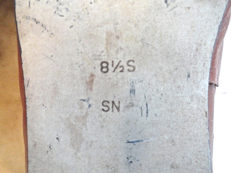 May include: Close-up of a shoe sole with the size marking "8 1/2 S" and the letters "SN" printed in black. The sole is a light beige colour with some wear and tear.