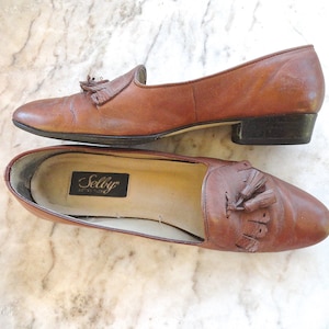 May include: A pair of brown leather loafers with tassel detailing. The shoes feature a low heel and a classic design. The interior of one shoe displays a "Selby" label. The shoes are on a marble surface.