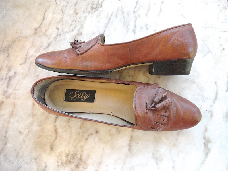 May include: A pair of brown leather loafers with tassel detailing. The shoes feature a low heel and a classic design. The interior of one shoe displays a "Selby" label. The shoes are on a marble surface.