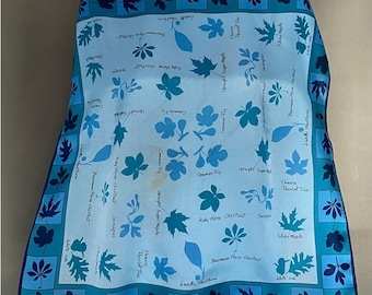 May include: A light blue scarf with a botanical leaf pattern. The scarf features various leaf illustrations in shades of blue, with the names of the leaves printed on the fabric. A dark blue border frames the scarf.