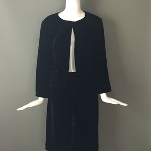 May include: A black velvet jacket with a long, flowing silhouette. The jacket has a simple, round neckline and a single button closure at the front. The sleeves are long and have a slight bell shape at the cuffs.