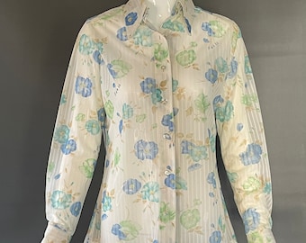 Puede incluir: Una camisa blanca de manga larga con botones y un estampado floral azul y verde. La camisa tiene cuello y una silueta ajustada.