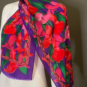 May include: A vibrant silk scarf featuring a floral pattern of red and pink calla lilies with green leaves against a purple background. The scarf has a fringed edge and a decorative border.