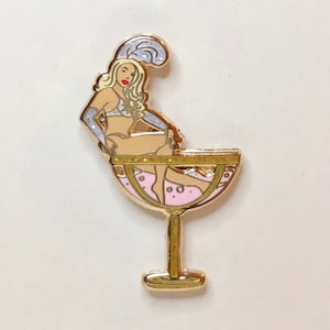 May include: A pin featuring a blonde showgirl in a cocktail glass. The pin has gold accents and a pink and white color scheme. The text "Adèle Wolf International Showgirl" and website address are also visible.