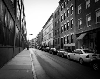 May include: A black and white photo of a city street with parked cars and buildings on either side. The street is empty except for a single person walking on the sidewalk.