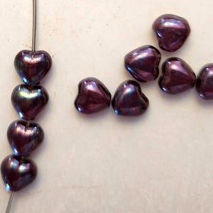 May include: Close-up of heart-shaped beads in a deep purple color with iridescent highlights. Some beads are strung on a wire, while others are scattered. The beads have a glossy finish and are arranged on a light-colored surface.