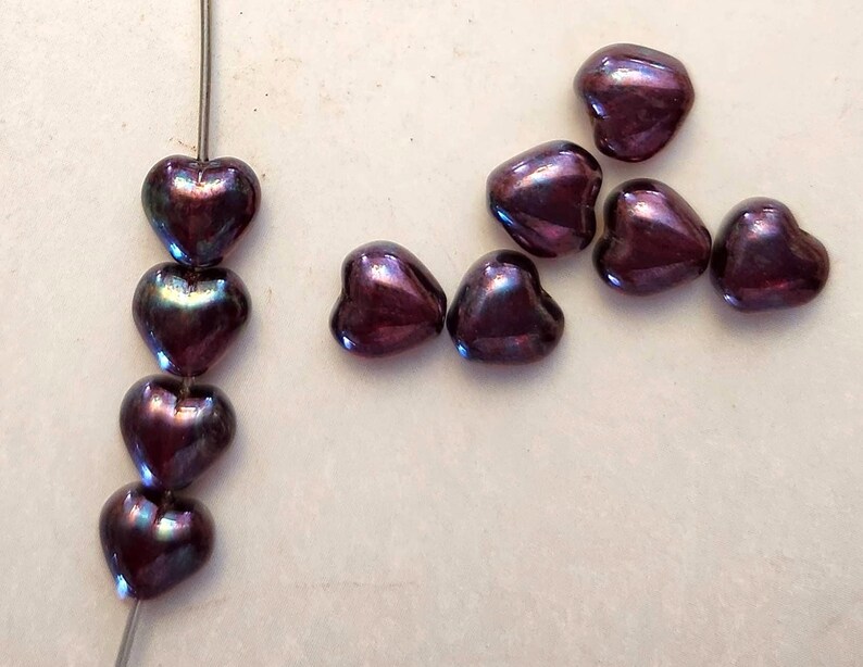 May include: Close-up of heart-shaped beads in a deep purple color with iridescent highlights. Some beads are strung on a wire, while others are scattered. The beads have a glossy finish and are arranged on a light-colored surface.
