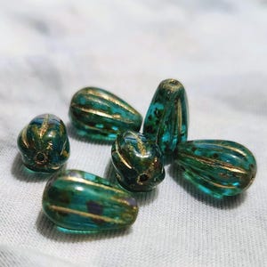 May include: Six teal and gold glass beads. The beads have a teardrop shape with gold stripes and accents. The beads are a mix of teal and green colors.
