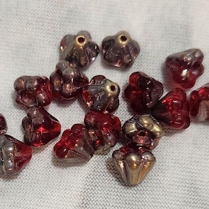 May include: A collection of red and gold-toned flower-shaped beads. The beads are scattered on a white fabric surface. The beads have a translucent quality, with a mix of red and gold colors. These beads are suitable for jewelry making and crafting.