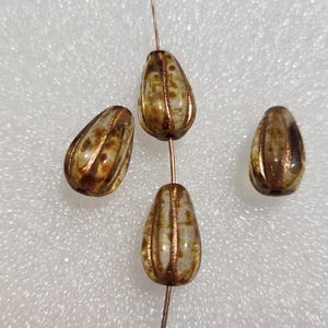May include: Four teardrop-shaped glass beads featuring a mottled brown and cream finish with vertical metallic copper-colored ridges. Two beads are strung on a thin wire, while two additional beads are positioned separately to the left and right.