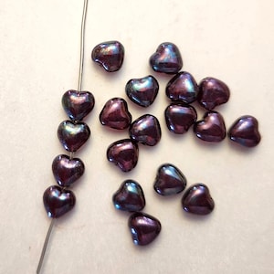 May include: A collection of iridescent heart-shaped beads in shades of purple, blue, and green. Some beads are strung on a silver wire, while others are scattered on a light-colored surface. The beads have a glossy finish.