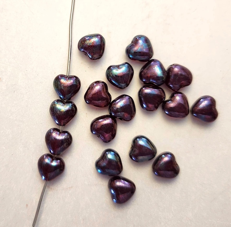 May include: A collection of iridescent heart-shaped beads in shades of purple, blue, and green. Some beads are strung on a silver wire, while others are scattered on a light-colored surface. The beads have a glossy finish.