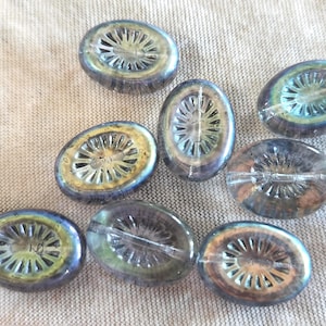 May include: A collection of iridescent oval-shaped glass beads. Each bead features a central, wheel-like design with radiating spokes. The beads have a mix of blue, green, and gold hues, creating a shimmering effect. They are arranged on a textured, neutral-colored surface.
