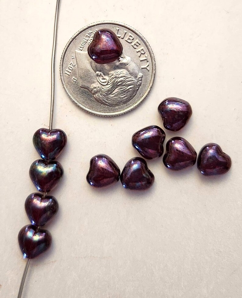 May include: A collection of heart-shaped beads in a deep purple color with iridescent highlights. Some beads are strung on a wire, while others are loose. A US dime is included for size reference. The beads have a glossy finish.