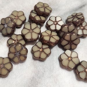 May include: A collection of flower-shaped beads. Each bead has a light beige center with a bronze-colored outline. The beads are scattered on a white fabric surface. These beads are ideal for jewelry making and crafting.