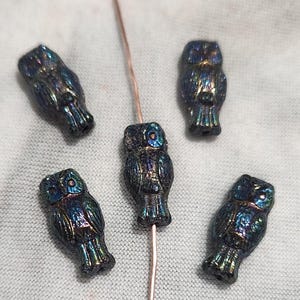 May include: Five iridescent owl-shaped beads. The beads are dark with a shimmering finish that reflects blues, greens, and purples. One bead is strung on a copper wire. The owls have detailed wings and facial features.
