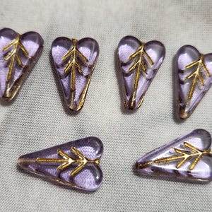 May include: Six translucent light purple glass beads shaped like hearts or leaves, each featuring a metallic gold-colored branch or arrow design embedded on the surface. The beads have a smooth, polished finish and a distinct triangular shape.