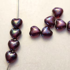 May include: Close-up of heart-shaped beads in shades of purple and iridescent green. Some beads are strung on a wire, while others are scattered. These beads are perfect for jewelry making and crafting projects.