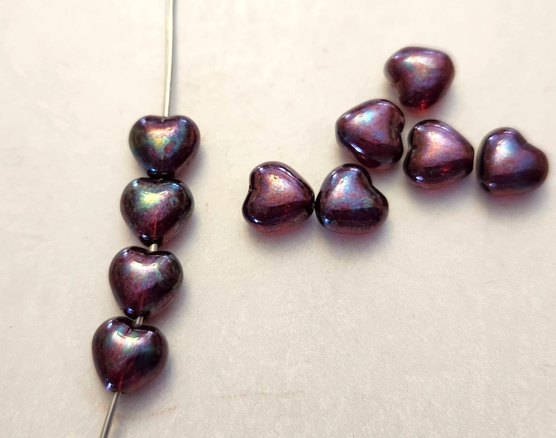 May include: Close-up of heart-shaped beads in shades of purple and iridescent green. Some beads are strung on a wire, while others are scattered. These beads are perfect for jewelry making and crafting projects.