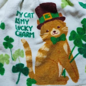 May include: White kitchen towel with a brown and orange cat wearing a green hat with a gold buckle. The cat has a green bow tie and the text "My cat is my lucky charm" is printed on the towel. The towel is decorated with green four-leaf clovers.
