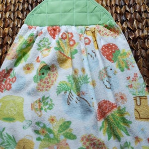 May include: A green tea towel with a floral pattern featuring strawberries, flowers, and gardening tools. The towel has a green quilted flap with a yellow button.