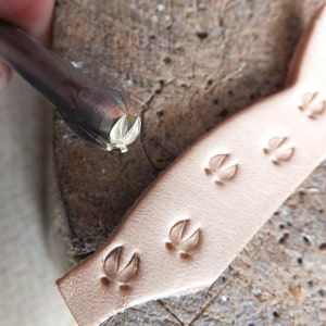 May include: Close-up of a leather craft tool with a paw print design. The tool is being used to stamp a leather strip with multiple paw prints.