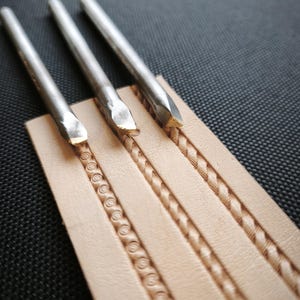 May include: Three metal leatherworking tools resting on a piece of tan leather with embossed designs. The leather has three different patterns: a series of circles, a braided design, and a textured line. The tools have silver handles and angled, gold-colored tips.