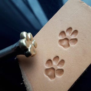 Może przedstawiać: Złoty metalowy stempel w kształcie odcisku łapy z drewnianym uchwytem. Stempel służy do tworzenia wzor&oacute;w odcisk&oacute;w łap na sk&oacute;rze.
