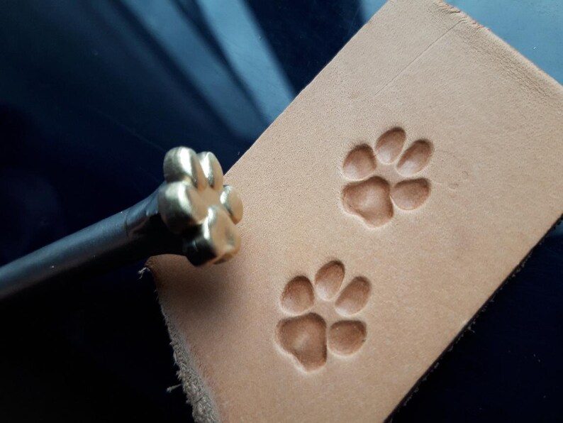 Może przedstawiać: Złoty metalowy stempel w kształcie odcisku łapy z drewnianym uchwytem. Stempel służy do tworzenia wzor&oacute;w odcisk&oacute;w łap na sk&oacute;rze.