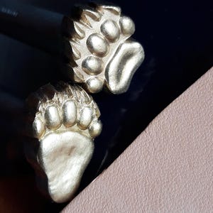 May include: Two gold-coloured metal bear paw stamps. The stamps are designed to be used for leather crafting.