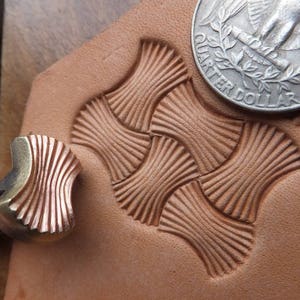 Può includere: Un primo piano di uno strumento per la stampa del cuoio con un design a forma di ventaglio. Lo strumento viene utilizzato per creare uno schema su un pezzo di cuoio. Una moneta da un quarto di dollaro americano è sullo sfondo per la scala.