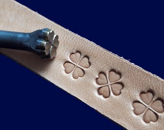 Op de afbeelding: Een close-up van een metalen gereedschap dat wordt gebruikt om leer te stempelen met een klaverbladontwerp. Het gereedschap wordt gebruikt om een ​​stuk leer te stempelen, en het klaverbladontwerp is zichtbaar op het leer.