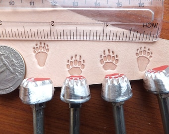 Op de afbeelding: Vier metalen berenpootstempels met rode rubberen inzetstukken. De stempels zijn gemonteerd op houten handvatten en worden gebruikt voor lederbewerking.