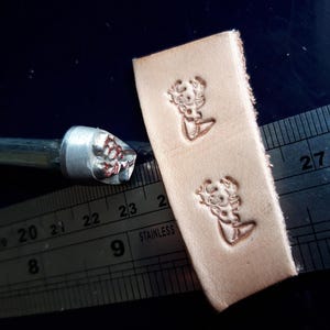 Może przedstawiać: Metalowy stempel do skóry ze szczegółowym wzorem, obok kawałka stemplowanej skóry. Skóra ma dwa odciski wzoru. Na zdjęciu znajduje się również metalowa linijka.