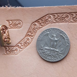 Könnte beinhalten: Ein Lederprägewerkzeug mit einem dekorativen Design. Das Werkzeug wird verwendet, um einen Rand auf einem Stück Leder zu erstellen. Eine US-amerikanische Viertel-Dollar-Münze dient als Größenvergleich.
