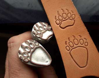 Op de afbeelding: Twee metalen berenpootstempels, een met een reliëf en de andere met een vlak oppervlak, worden getoond naast een stuk bruin leer met twee ingegraveerde berenpootontwerpen.