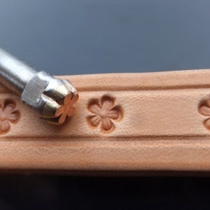 Puede incluir: Una herramienta de estampado de cuero con un diseño de flor de cinco puntas. La herramienta está apoyada sobre un trozo de cuero con varias impresiones del diseño de la flor.