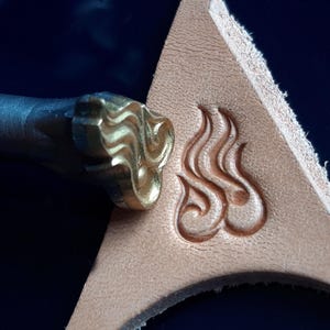Peut inclure: Un outil de marquage en cuir avec un motif doré. L'outil est utilisé pour créer un motif décoratif sur un morceau de cuir.