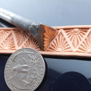 May include: A close-up of a leather stamping tool with a sunburst design. The tool is being used to create a pattern on a piece of leather. A US quarter is included for scale.