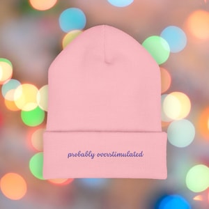 May include: A pale pink beanie hat with the embroidered phrase "probably overstimulated" in dark blue. The hat has a folded brim and is made of a soft fabric. The background is a blur of colourful bokeh lights.