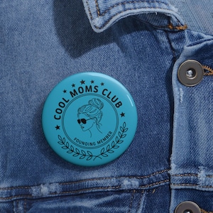 Op de afbeelding: Een ronde, turquoise button met de tekst "COOL MOMS CLUB" in een cirkelvormig ontwerp. De button toont een silhouet van een vrouw met een zonnebril, omringd door sterren en een lauwerkrans. De woorden "FOUNDING MEMBER" zijn ook opgenomen.