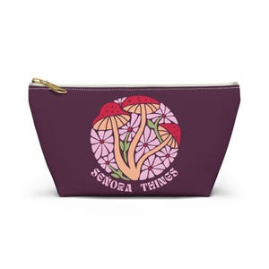 Puede incluir: Una bolsa con cremallera de color ciruela con un tirador dorado. La bolsa presenta un diseño circular de setas y flores en tonos rojos, rosas y beige, con el texto "Senora Things" en una fuente curva.