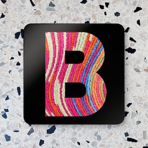 Puede incluir: Un posavasos cuadrado negro con una gran letra "B". La letra está rellena con un patrón colorido y texturizado de rayas rosas, naranjas, azules y verdes. El posavasos tiene esquinas redondeadas.