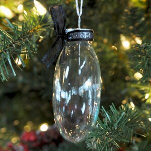 May include: A clear, oval-shaped glass ornament with a hollow center. The top features a silver-colored metal cap wrapped in a black ribbon tied into a small bow. A thin white string is attached to the top for hanging the decorative piece.