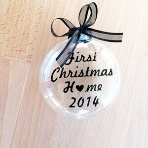 May include: A clear, spherical glass bauble featuring a black ribbon bow at the top. The text "First Christmas Home" is printed in black script, with a small black heart replacing the letter "o" in "Home," followed by the year "2014" below.